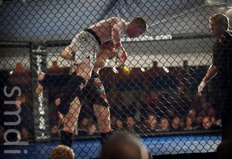 Man lifting another man in an MMA ring surrounded by chain link fencing